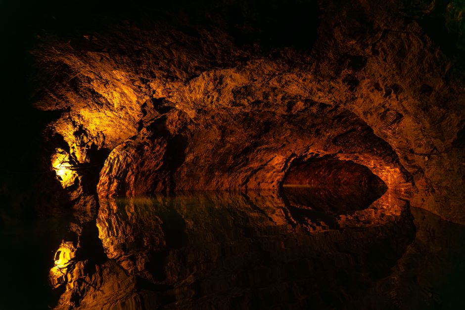 An underground cave interior with rough, natural rock formations illuminated by soft, warm artificial lighting that highlights the textures and layers of the stone. The cave features a narrow, winding