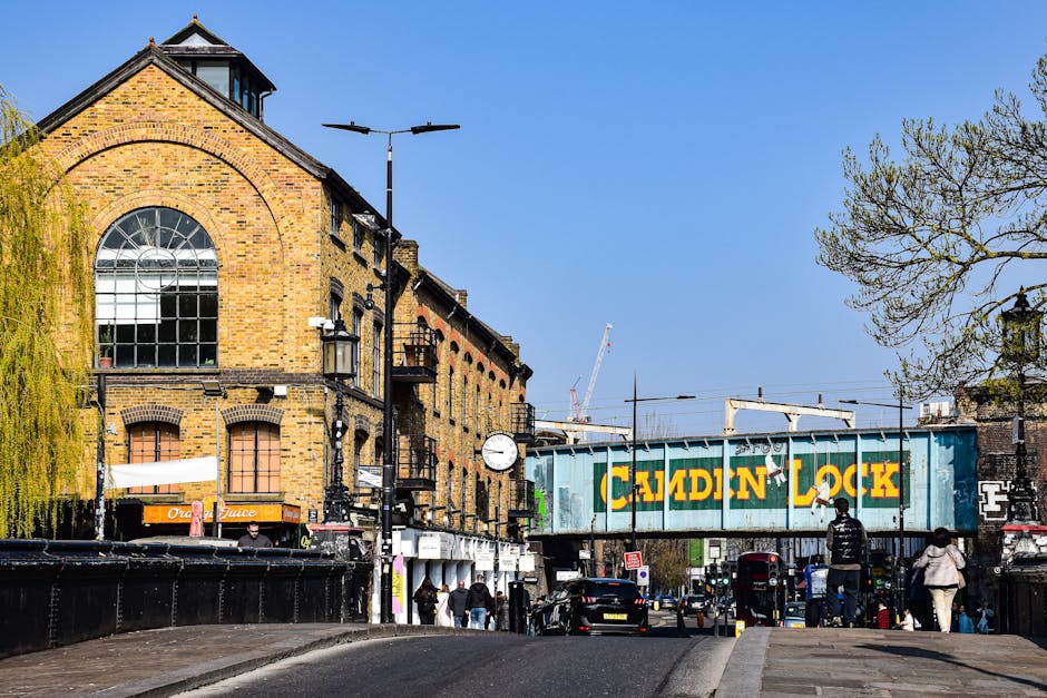 The image depicts a busy loading bay area outside a historic brick building with large arched windows, situated under a bright blue sky. Several pedestrians are walking along the pavement and near the