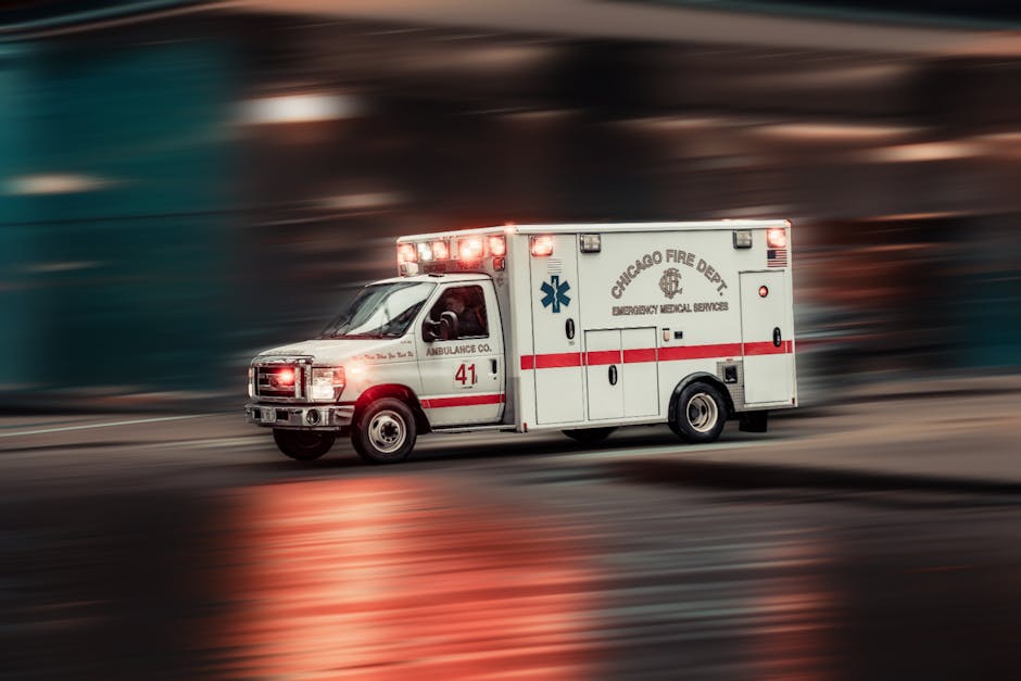 A white ambulance vehicle with red and blue markings is moving at speed on a city street during low light conditions, with its emergency lights flashing. The vehicle belongs to the Chicago Fire Depart